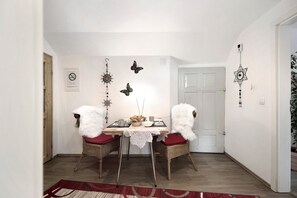 Un coin repas avec une table dressée pour deux personnes, deux chaises en osier avec des coussins rouges, un tapis à motifs rouges et blancs, et des décorations murales comprenant des papillons et un ornement en forme d’étoile.