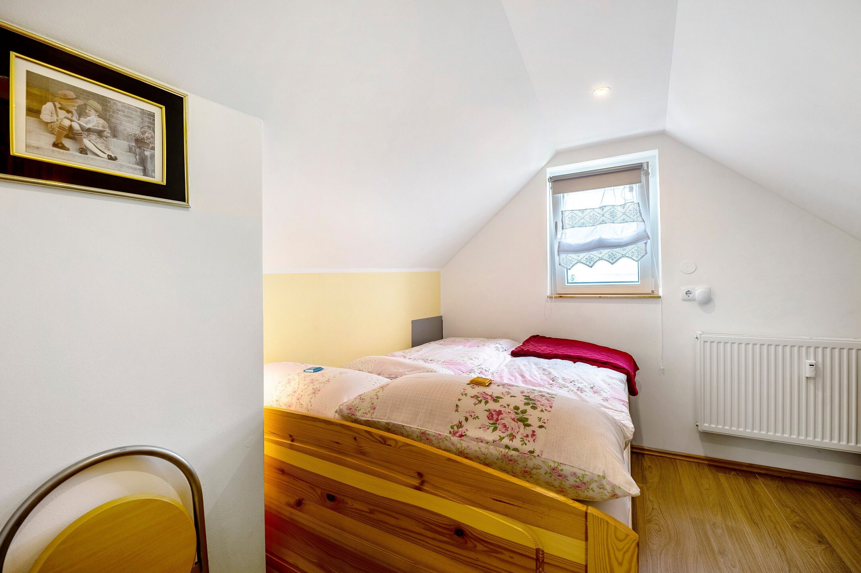 Une chambre avec un lit simple, une fenêtre avec des rideaux, un radiateur et un tableau encadré au mur.