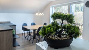 Un arbre bonsai en pot est posé sur une table blanche dans un coin repas moderne, avec un plancher en bois, des chaises noires et une grande fenêtre.
