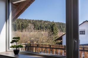 Une fenêtre donnant sur une colline boisée, une clôture en bois et un arbre bonsai en pot placé sur le rebord de la fenêtre.
