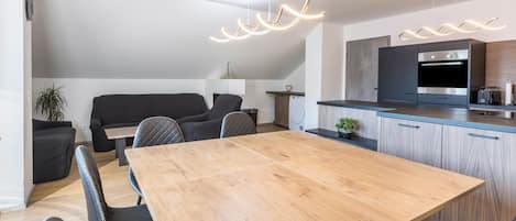 A modern kitchen with a built-in oven, a dining area with a wooden table and black chairs, and a black sofa in a living area.