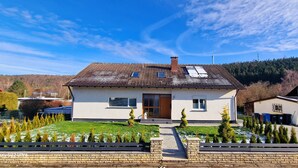 Une maison à deux étages, avec un toit en tuiles, une porte d’entrée et un petit jardin planté de jeunes arbres.