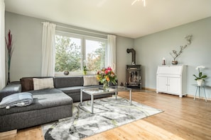 Un salon moderne avec un canapé d’angle gris, une table basse en verre, une cheminée et une vue sur les arbres à travers de grandes fenêtres.