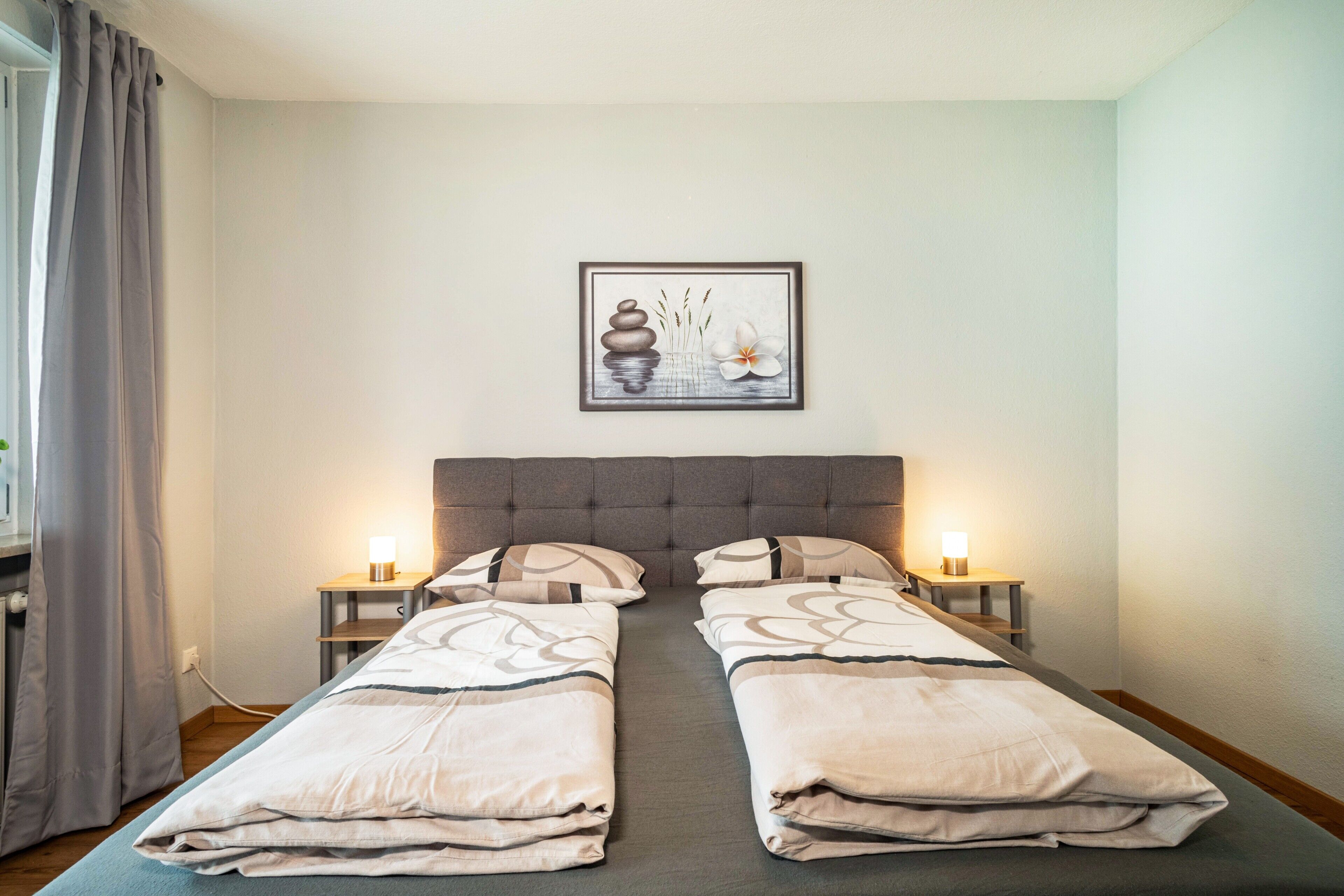 A double bed with a grey headboard, two bedside tables with lamps, a framed artwork on the wall, and a window with curtains.