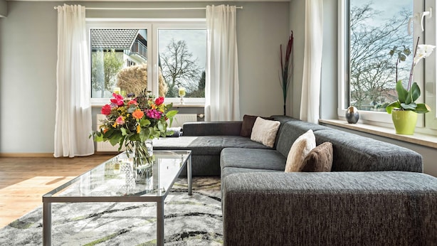 Ein modernes Wohnzimmer mit Ecksofa, Glastisch und Blick auf ein Gebäude durch große Fenster.