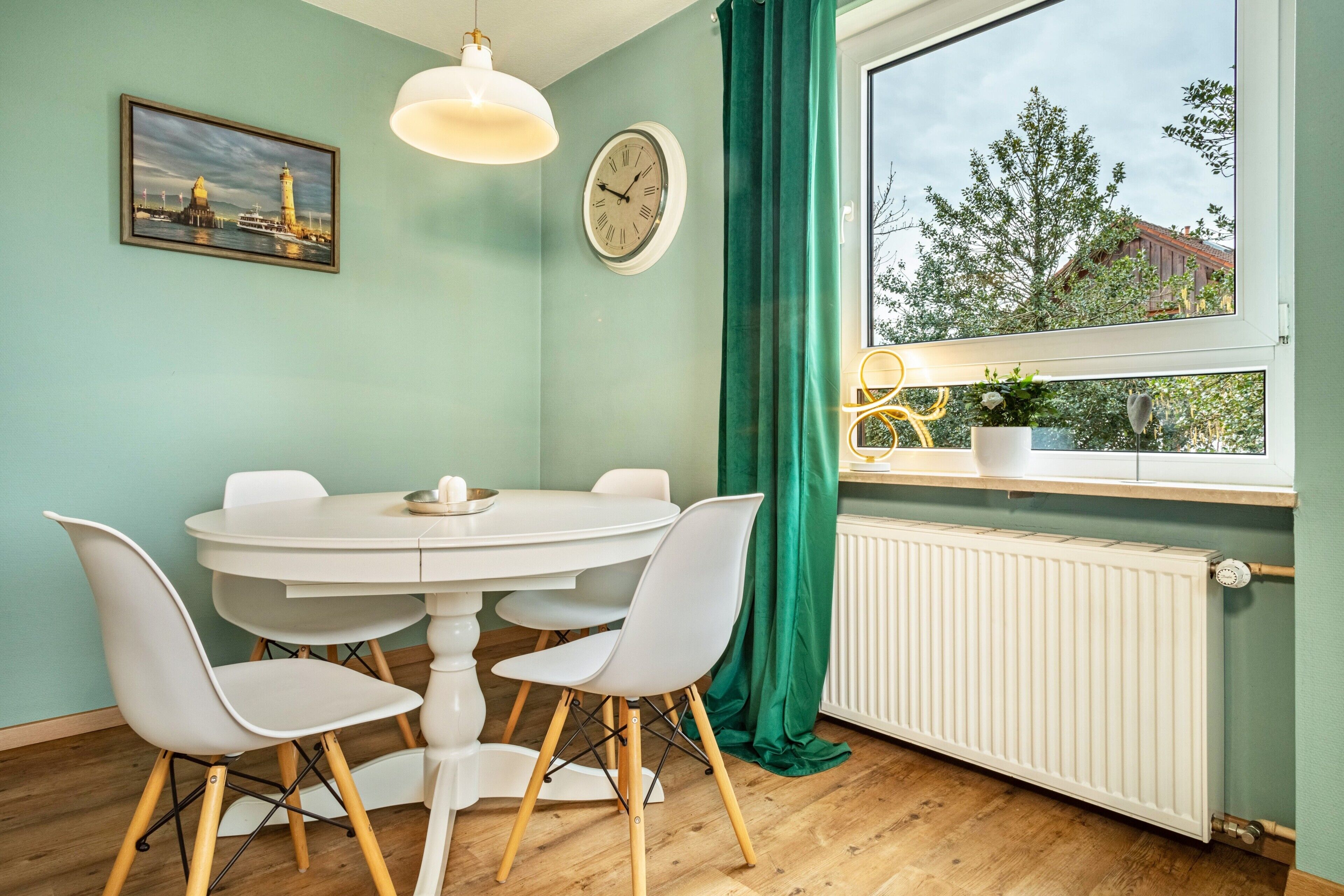 A dining area with a round table, four chairs, a clock, a window with a view of trees and buildings, and a radiator.