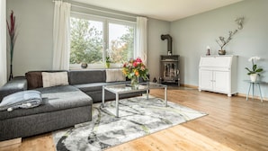 Ein modernes Wohnzimmer mit einem grauen Ecksofa, einem Glastisch, Holzboden, einem Kamin und Blick auf Bäume durch große Fenster.