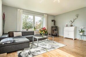 Una sala de estar moderna con un sofá seccional gris, una mesa de centro de vidrio, piso de madera, chimenea y vista a árboles a través de amplias ventanas.