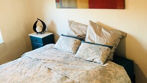 Quarto com uma cama grande, duas almofadas, uma mesa de cabeceira, um objeto decorativo e uma pintura abstrata colorida na parede.