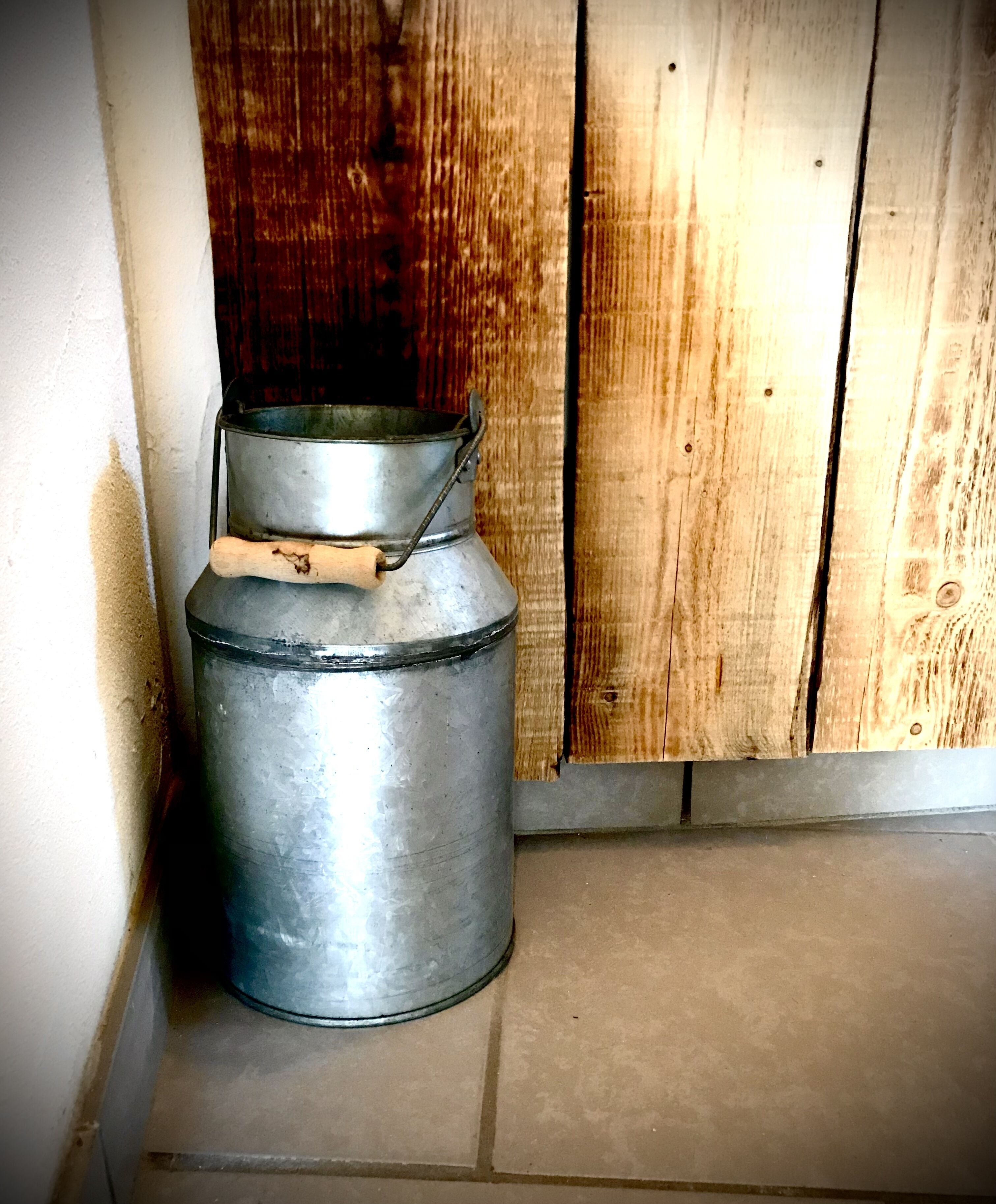 A metal container with a lid and handle, placed against a wooden wall and a tiled floor.