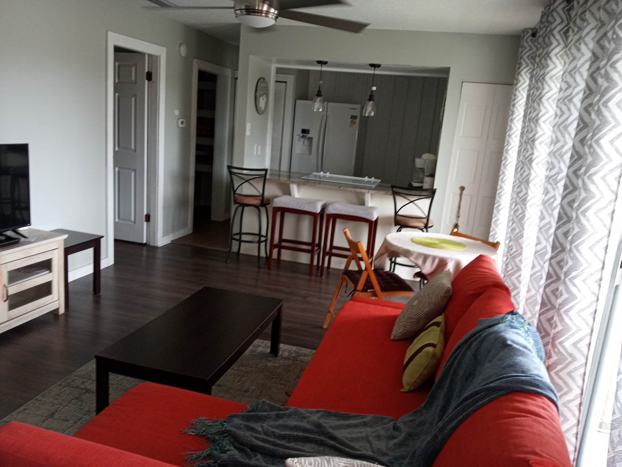 Ein Wohnzimmer mit einem roten Sofa, einem schwarzen Couchtisch und einem Fernseher auf einer Holzkommode. Es gibt eine Essecke mit einem weißen Tisch und Stühlen, und im Hintergrund befindet sich eine Küche.