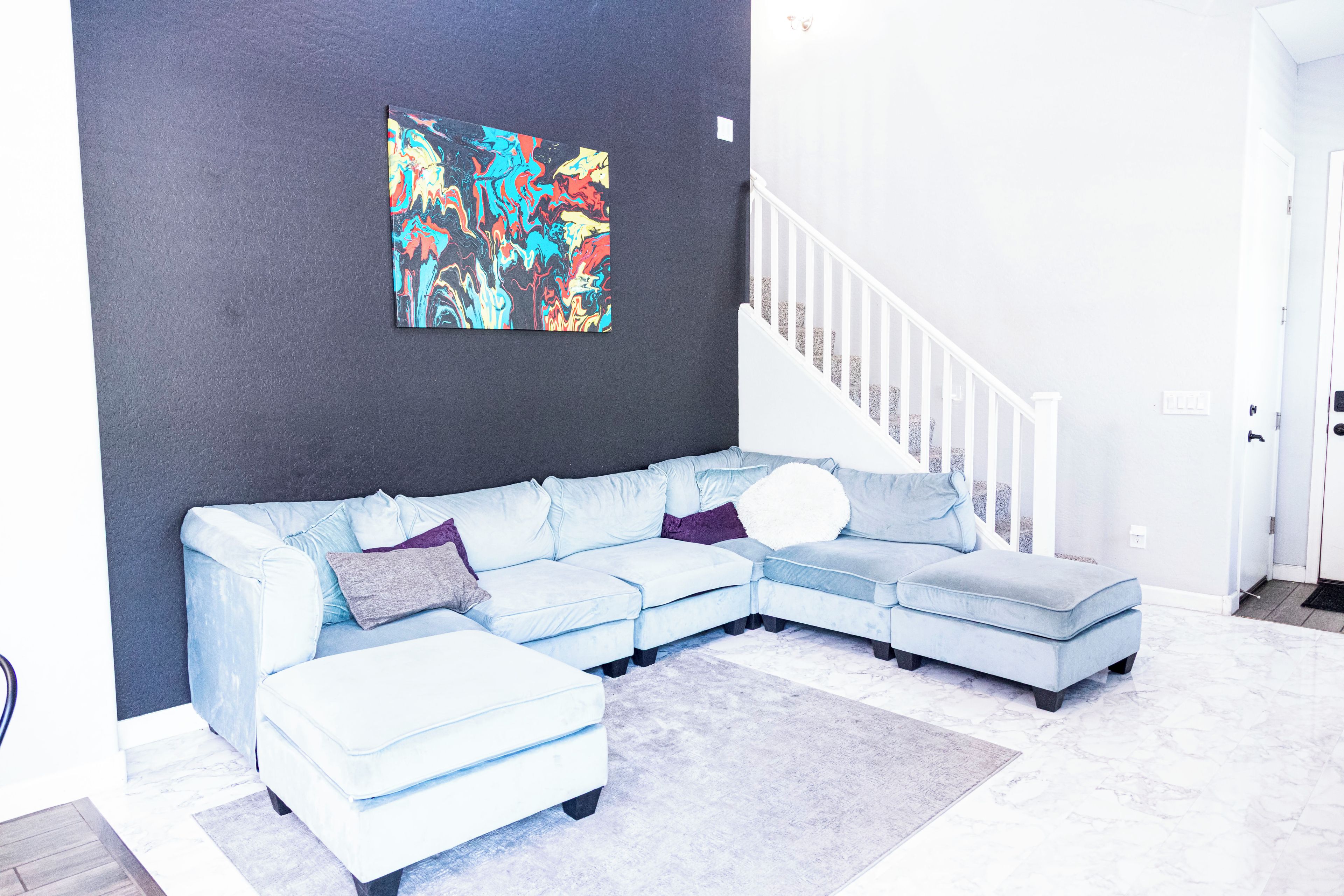 A modern living room with a light blue sectional sofa, a white round throw pillow, a colorful abstract painting on the wall, and a white staircase.