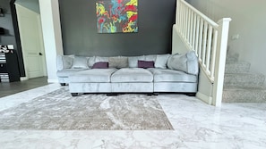 A living room with a grey sectional sofa, a patterned rug, a marble floor, a staircase, and a colorful abstract painting on the wall.