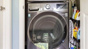 A laundry room with two stacked front-loading washing machines, a shelf with cleaning supplies, and a door leading to another room. - Luxury Gated Retreat – Resort Amenities, Pool, Spa, Gym (Mesa)