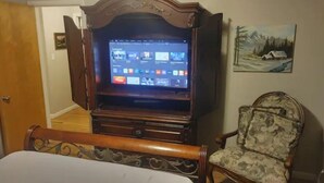 A room with a wooden entertainment center housing a flat-screen TV displaying a media library. A patterned armchair is positioned to the right, and a bed with a white headboard is in the foreground. - Self Check-In & Free Wi-Fi in this Fresh Remodel! (Idaho Falls)