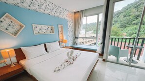 A hotel room with a large bed, a bedside table with a lamp, a window with a view of greenery, and a balcony with a chair. - Natalie House Kata Beach (Karon)