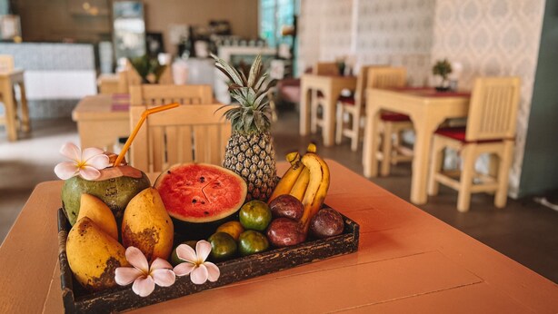A fruit platter on a table in a restaurant with wooden chairs and a patterned wall in the background. - Natalie House Kata Beach (Karon)