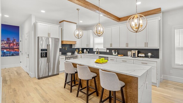 A modern kitchen with white cabinets, a stainless steel refrigerator, a marble island with bar stools, and pendant lighting.
