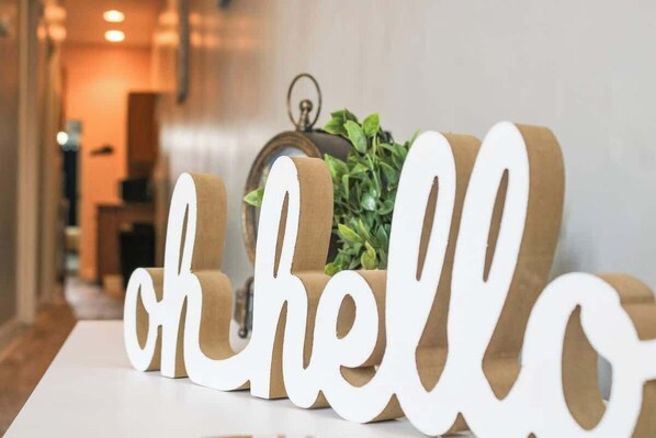 A white sign with the words 'oh hello' in a cursive font, placed on a white surface with a plant and a lantern in the background.