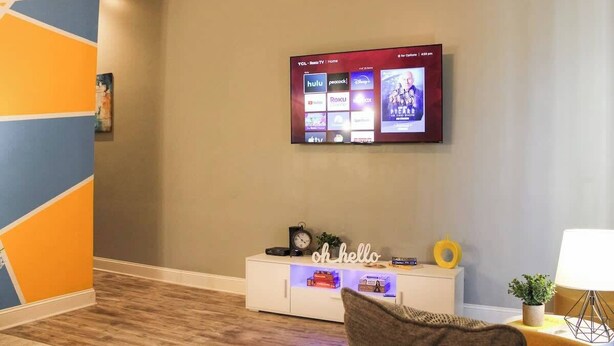 Una sala de estar con un televisor de pantalla montado mostrando servicios de transmisión, un aparador blanco con objetos decorativos y una lámpara moderna.