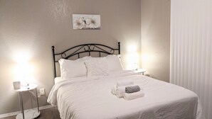 Ein ordentlich bezogenes Bett mit weißen Leinen, zwei Nachttischlampen, ein kleiner Nachttisch und ein an der Wand hängendes Bild mit Blumen.
