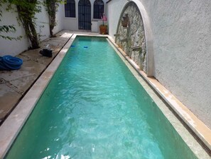 Una piscina rectangular con agua cristalina, rodeada por un muro de piedra y un edificio blanco con ventanas de arco.