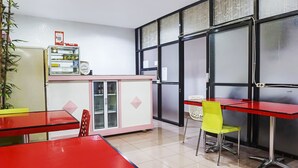 A compact kitchen with a refrigerator, a small pantry, and a window with a view. There are two red tables with chairs, one of which has a yellow chair. - Hotel O Homestay Madu Makassar (Makassar)
