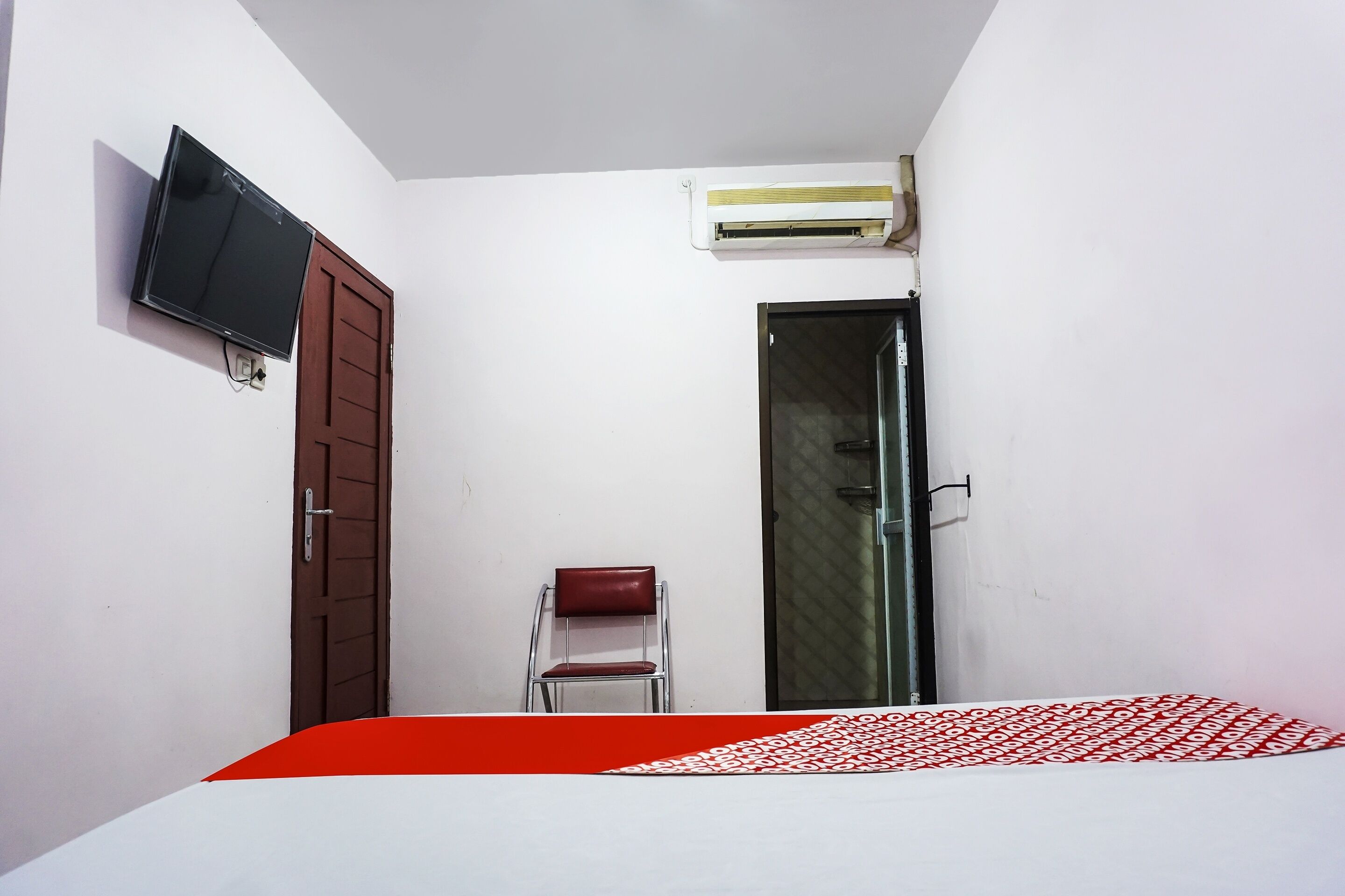 A small, simple room with a bed, a TV, an air conditioner, and a door.