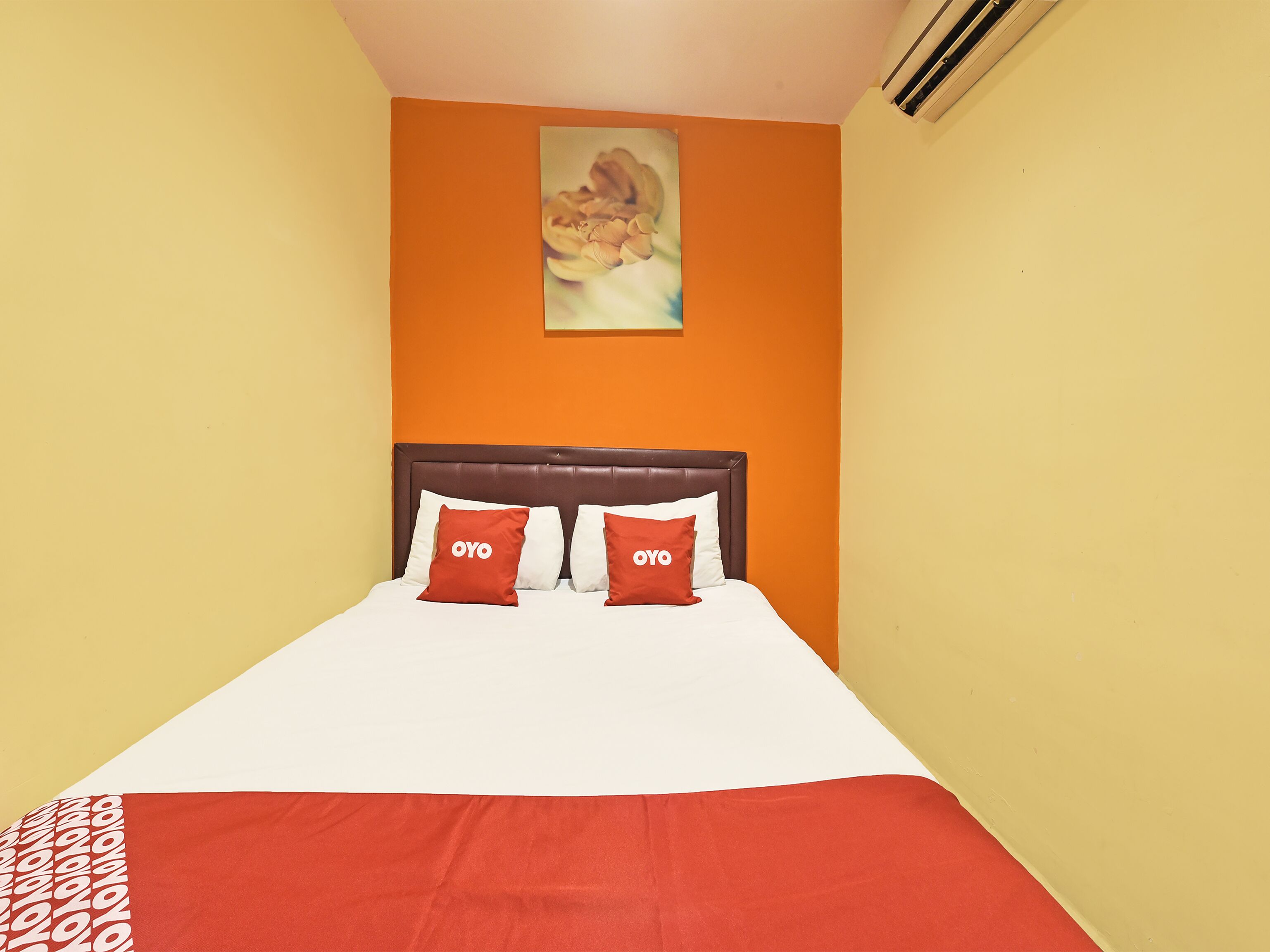 A hotel room with a bed, two pillows with "OYO" branding, an orange wall, and a painting above the bed.