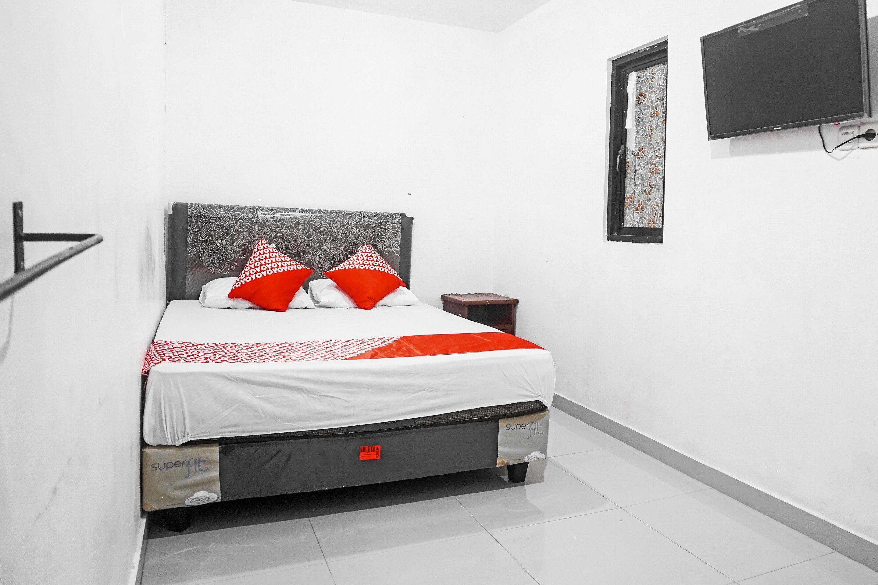 A bed with a white and red bedspread, two red pillows, and a grey headboard. There is a small wooden nightstand beside the bed.