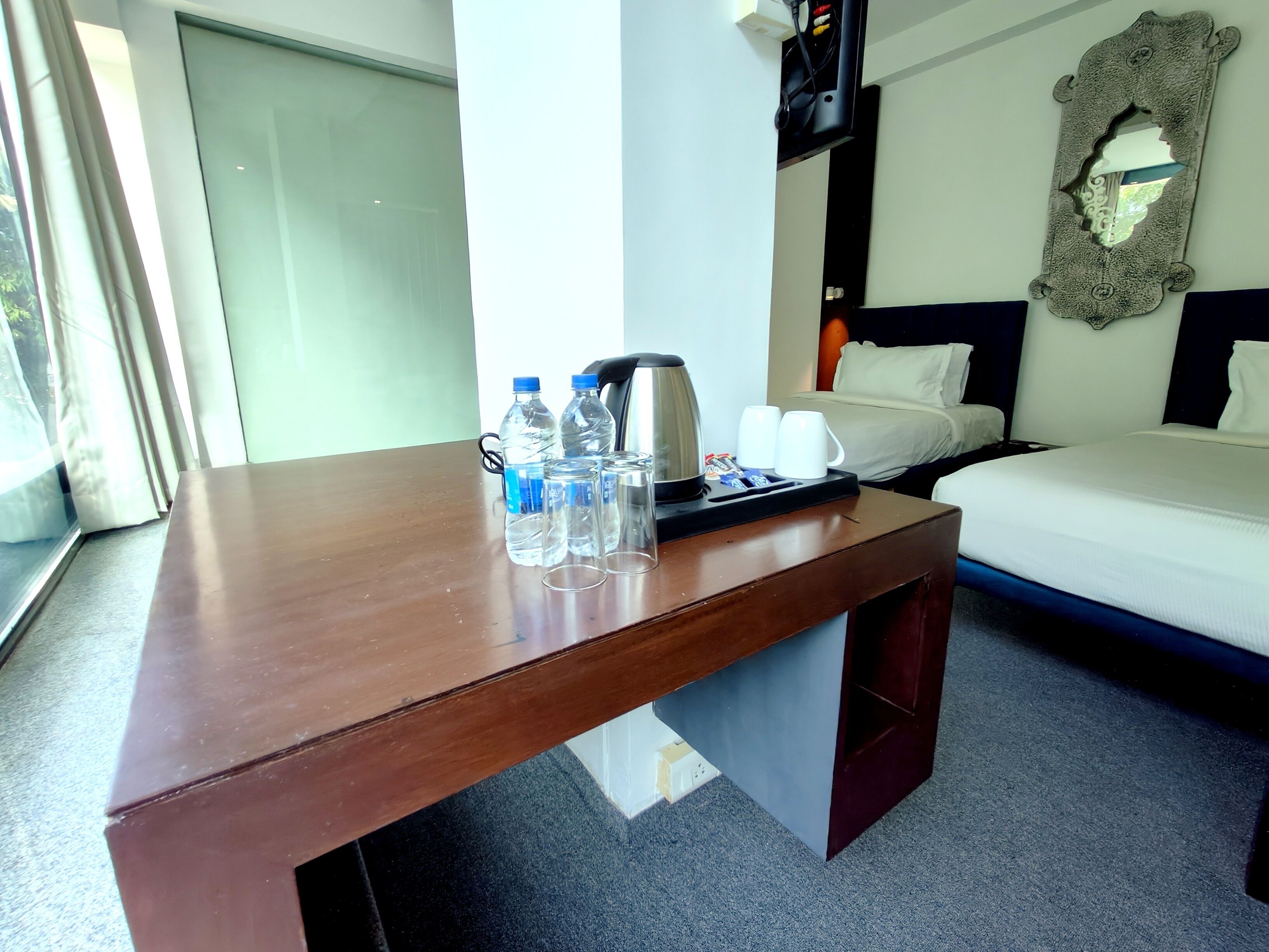 Une chambre d’hôtel avec un bureau en bois, un lit, une table de chevet avec une bouilloire, des tasses et des bouteilles d’eau.