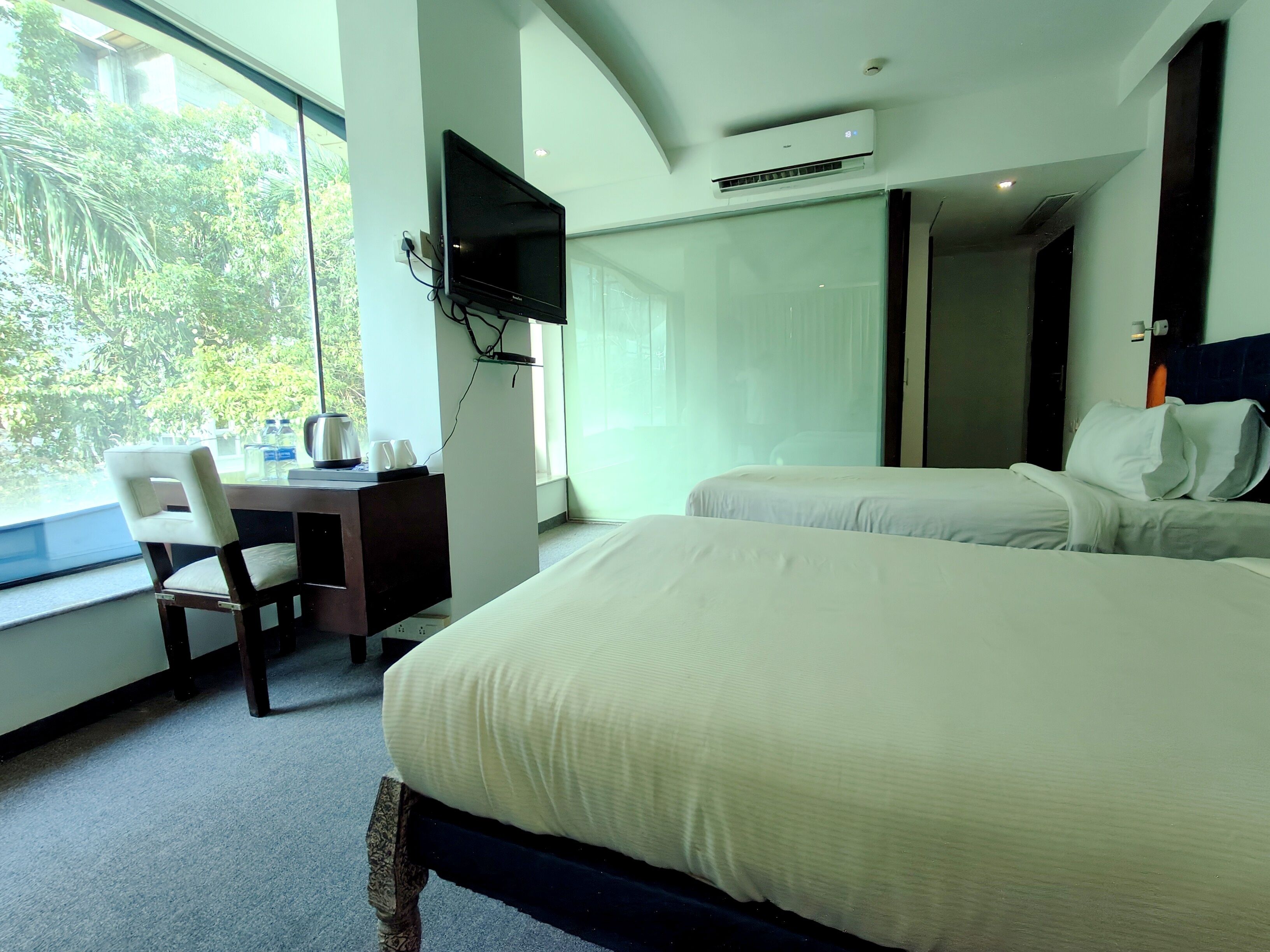 A hotel room with two beds, a desk with a chair, a TV mounted on the wall, and a large window offering a view of greenery outside.