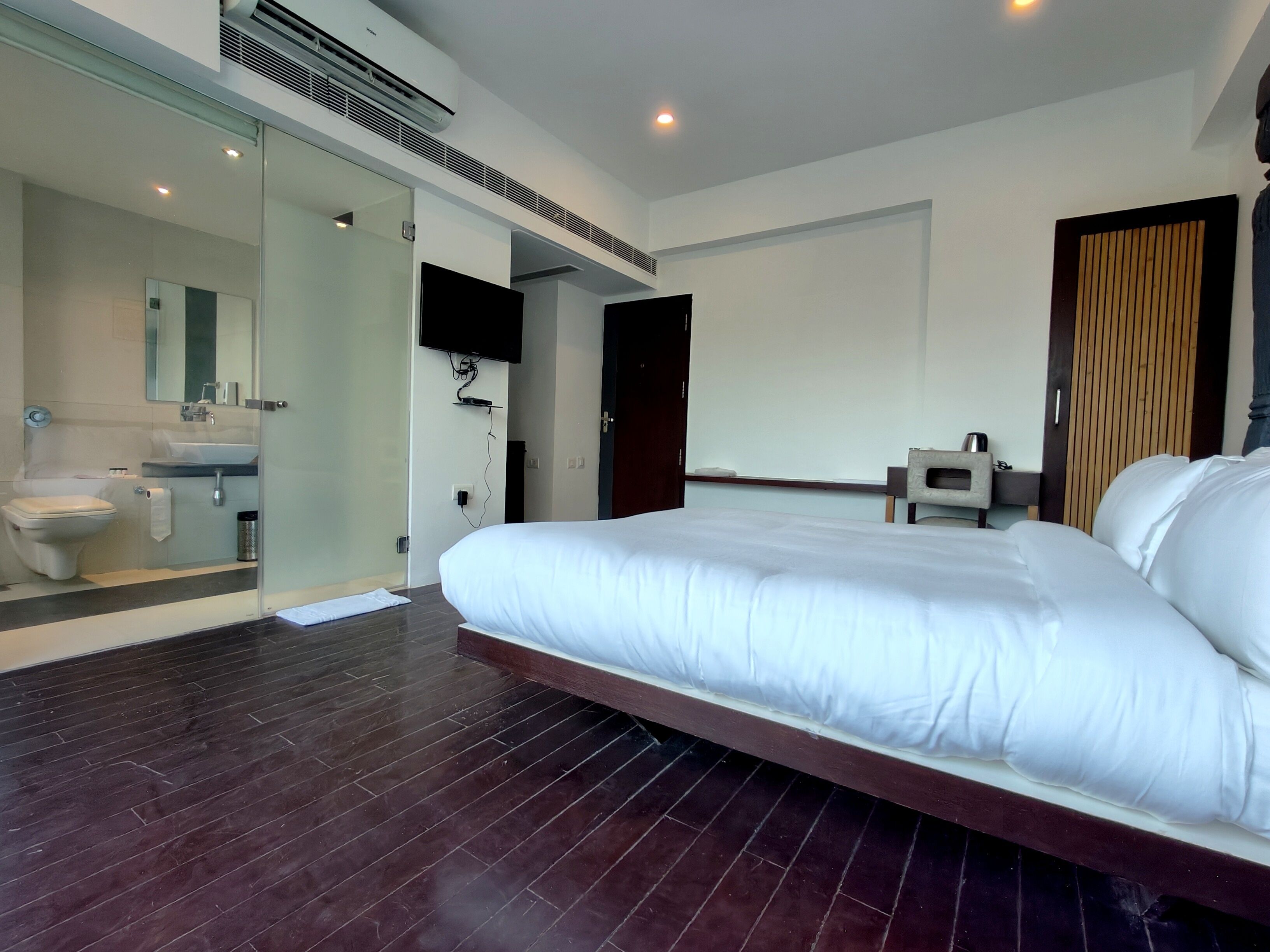 A modern hotel room with a large bed, a separate bathroom with a toilet and sink, a flat-screen TV mounted on the wall, and a wooden door.