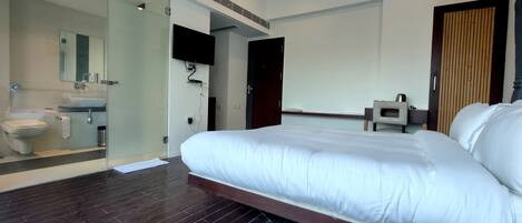 A modern hotel room with a large bed, a separate bathroom with a toilet and sink, a flat-screen TV mounted on the wall, and a wooden door.