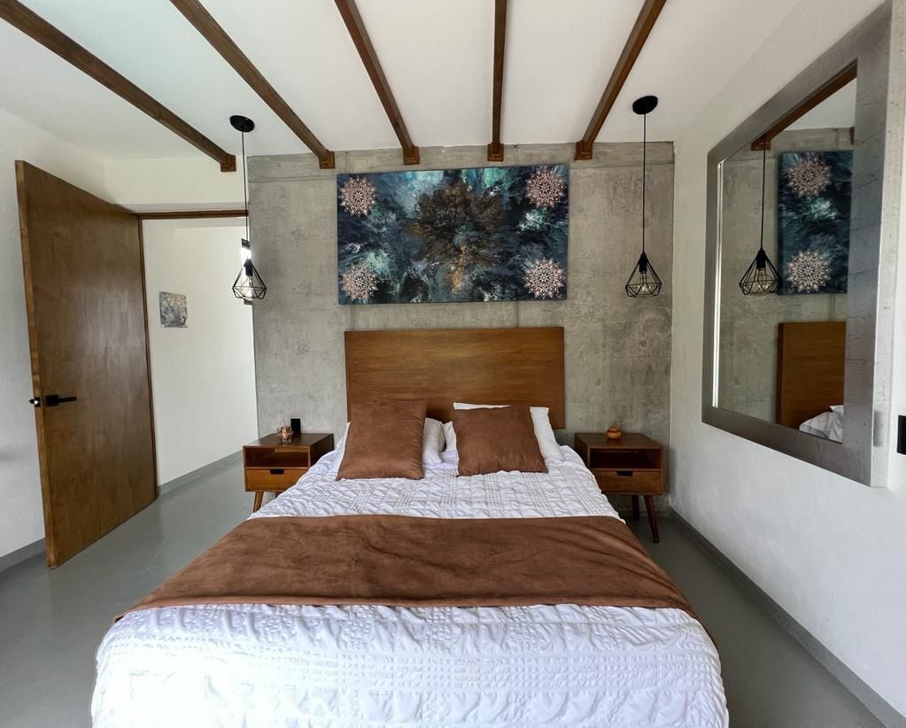 A bedroom with a large bed, wooden headboard, two bedside tables, a nightstand, a large mirror, and abstract wall art.