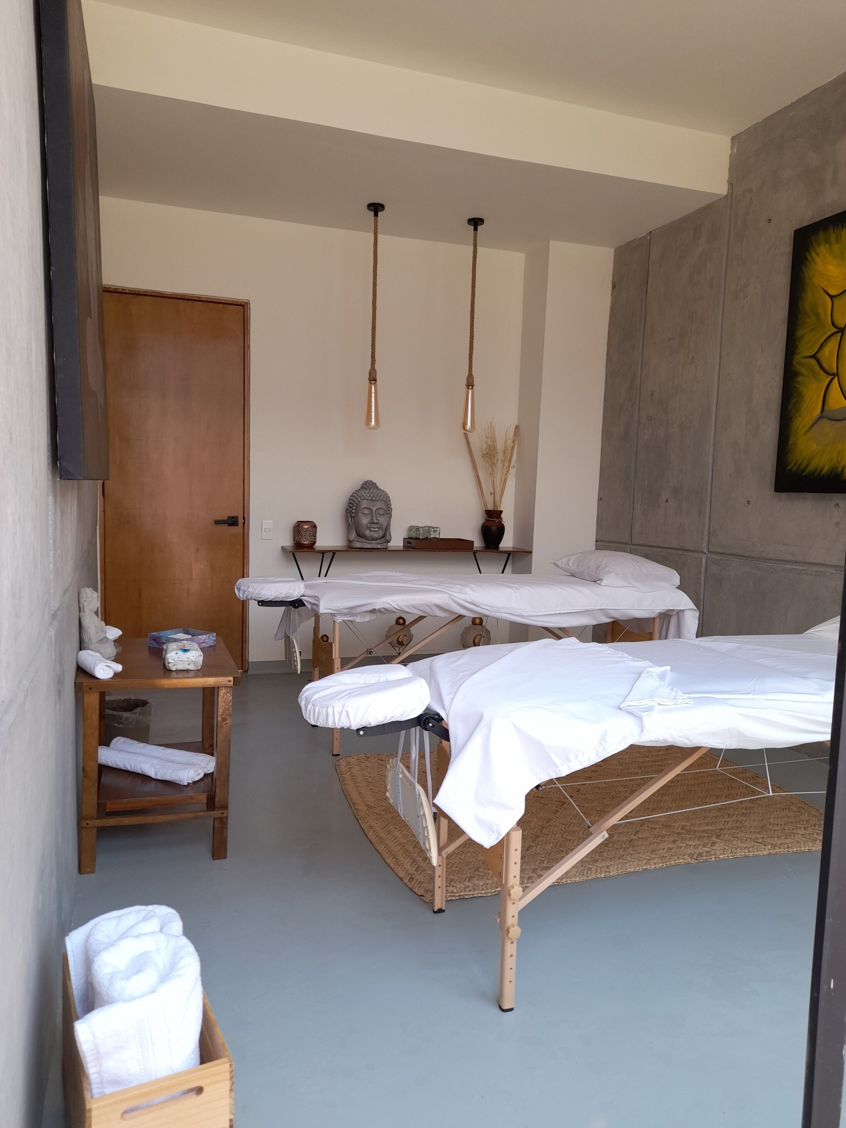 Una habitación con dos mesas de masaje, un banco de madera con toallas, un estante con objetos y un cuadro colgado en la pared.