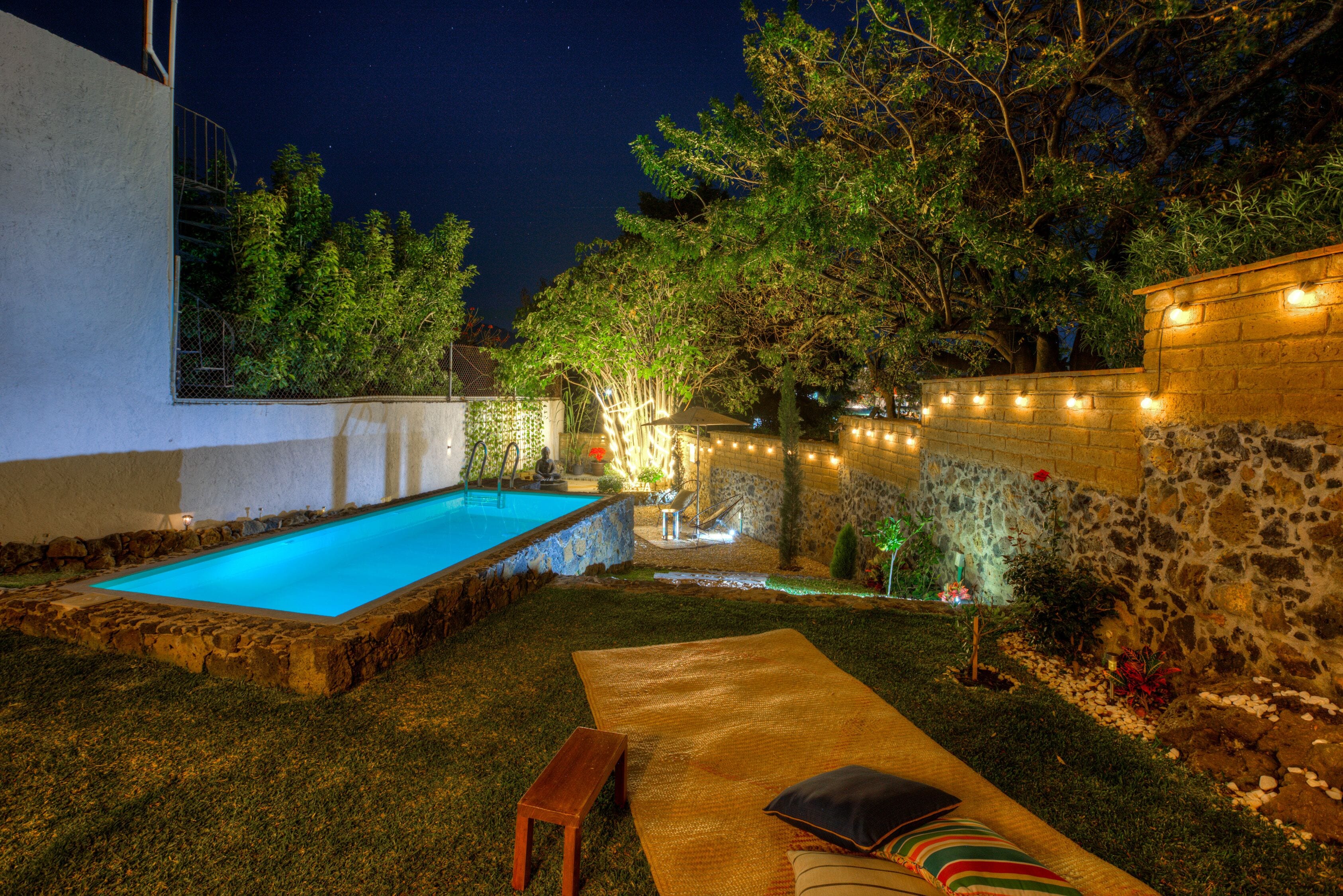 Piscina con bordo in pietra, zona relax con panche e cuscini, e luci decorative lungo un muro in pietra.
