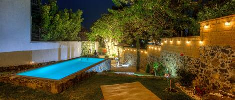 A swimming pool with a stone border, a lounge area with a bench and cushions, and string lights along a stone wall.