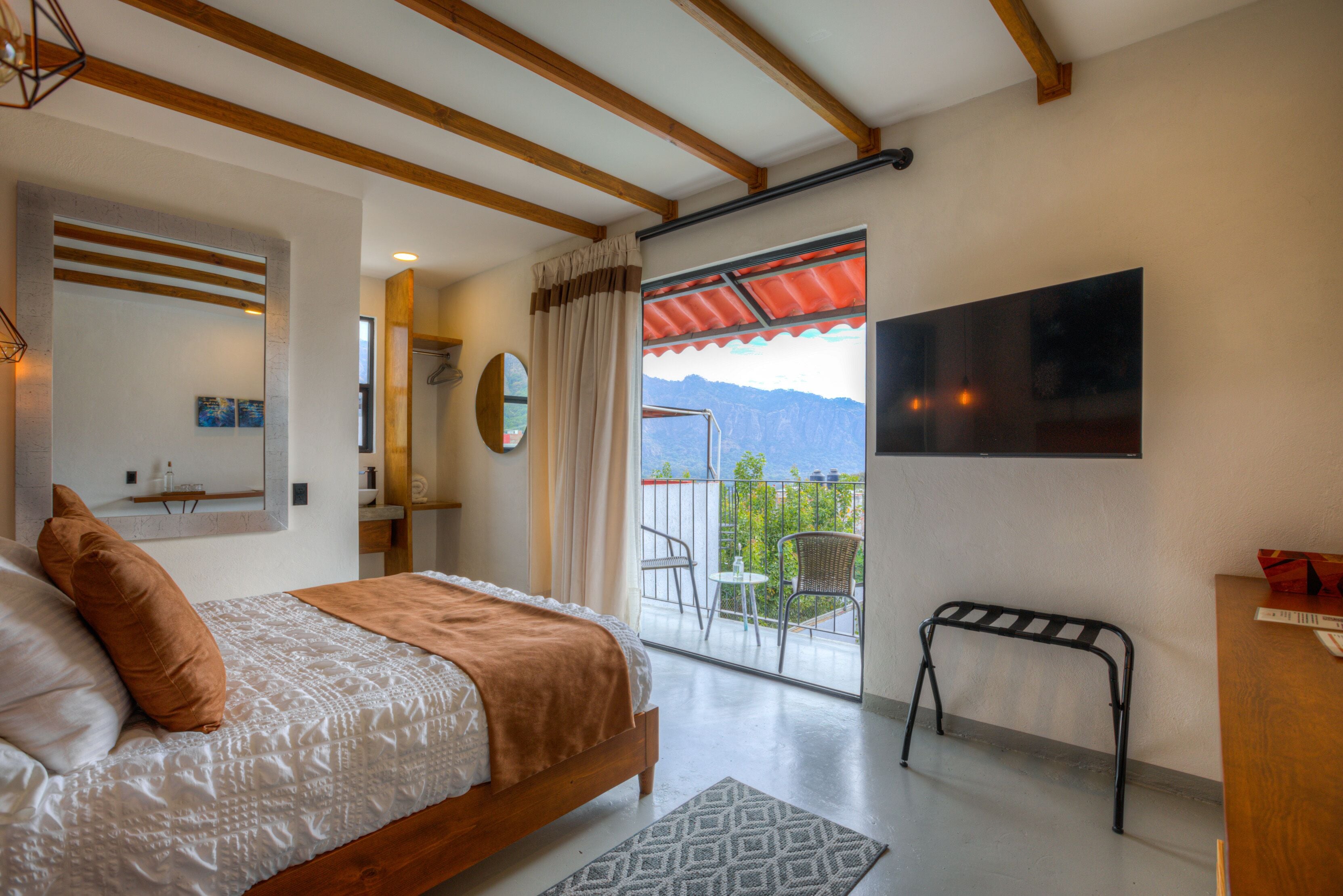 Un dormitorio con cama, mesita de noche, espejo, ventana con cortinas, balcón con sillas y televisor en la pared.