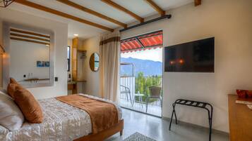 Un dormitorio con cama, mesita de noche, espejo, ventana con cortinas, balcón con sillas y televisor en la pared.