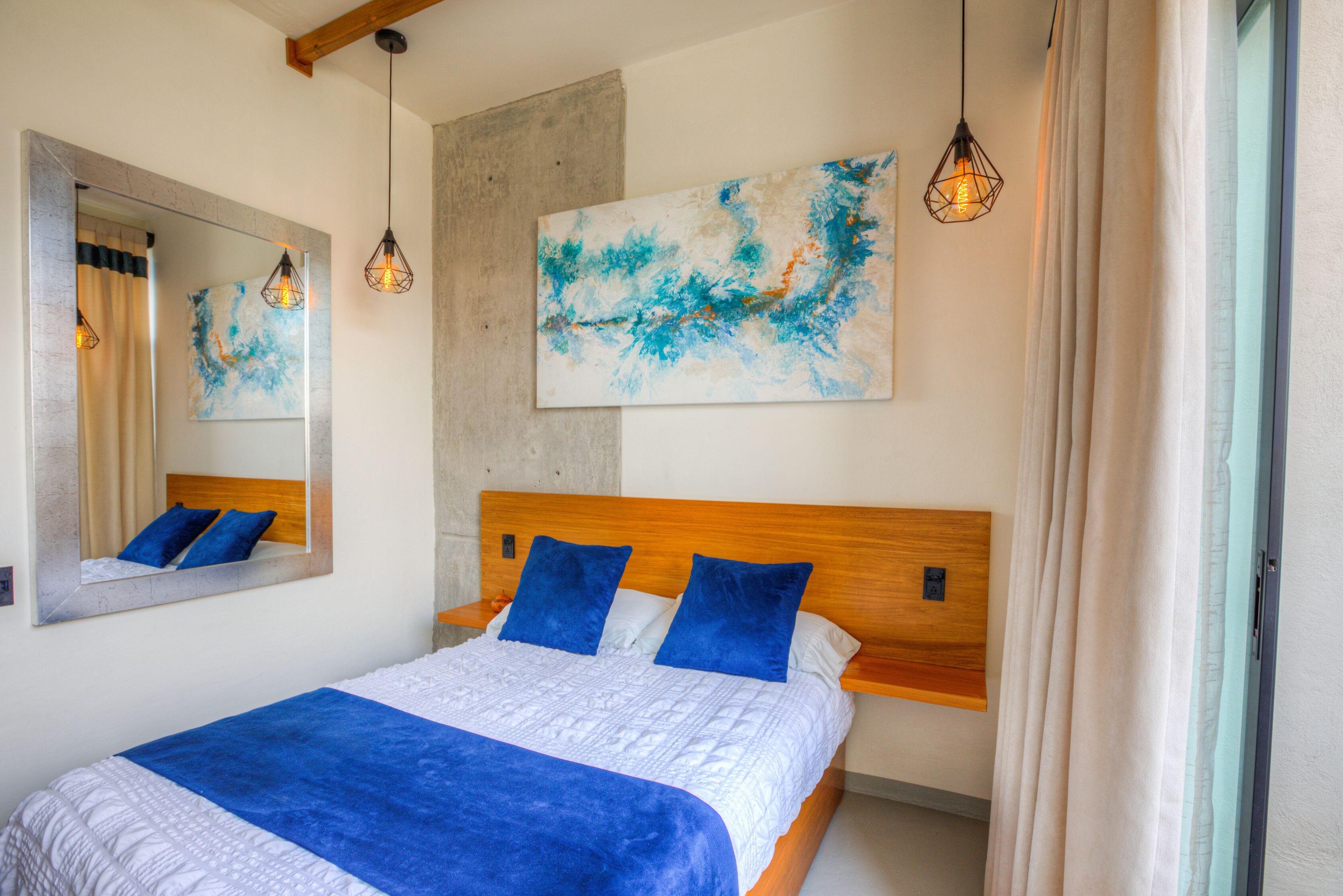 A neatly made bed with a blue blanket and pillows, a large abstract painting on the wall, and a wooden headboard.