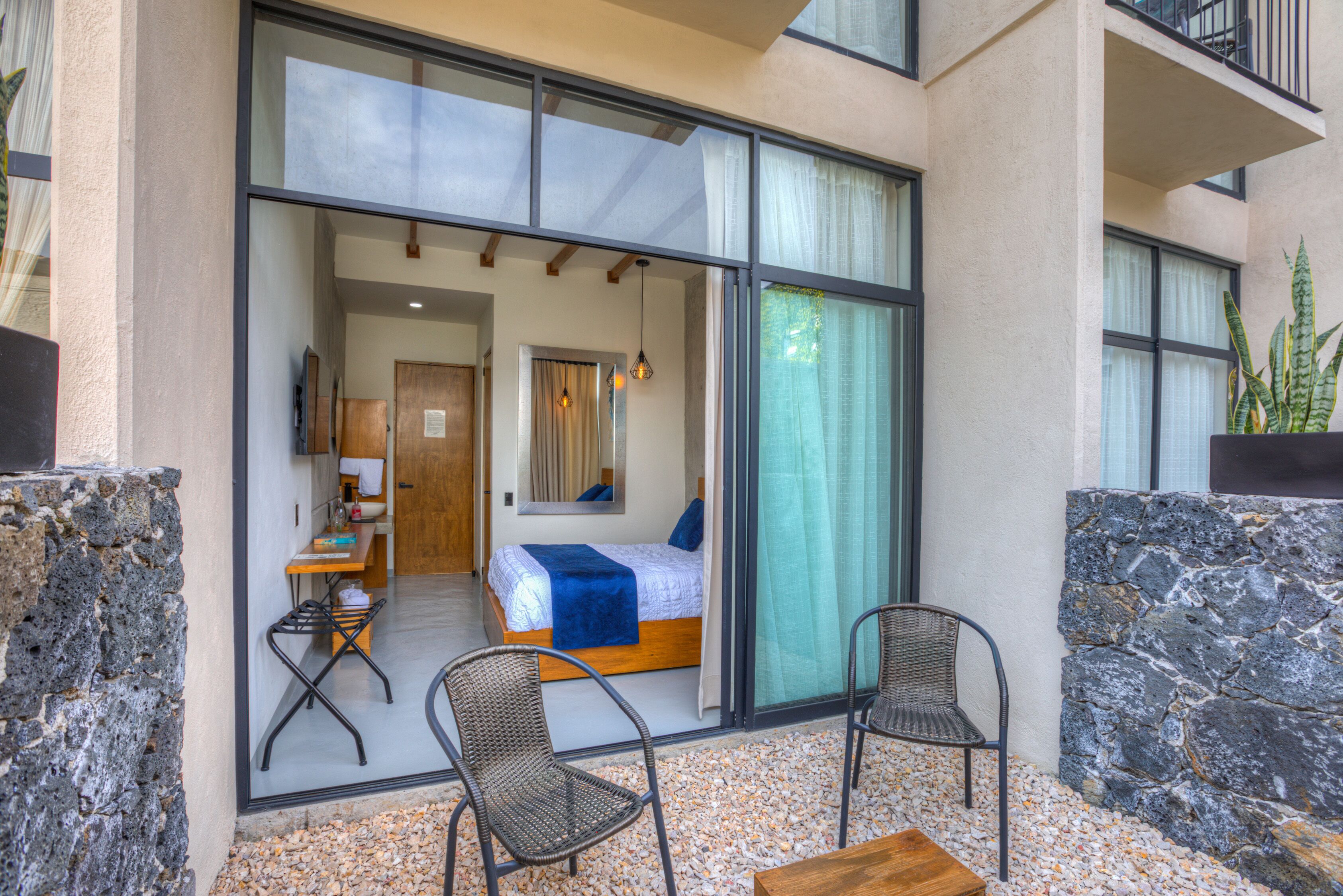 A modern hotel room with a large sliding glass door leading to a balcony with two wicker chairs and a wooden table.