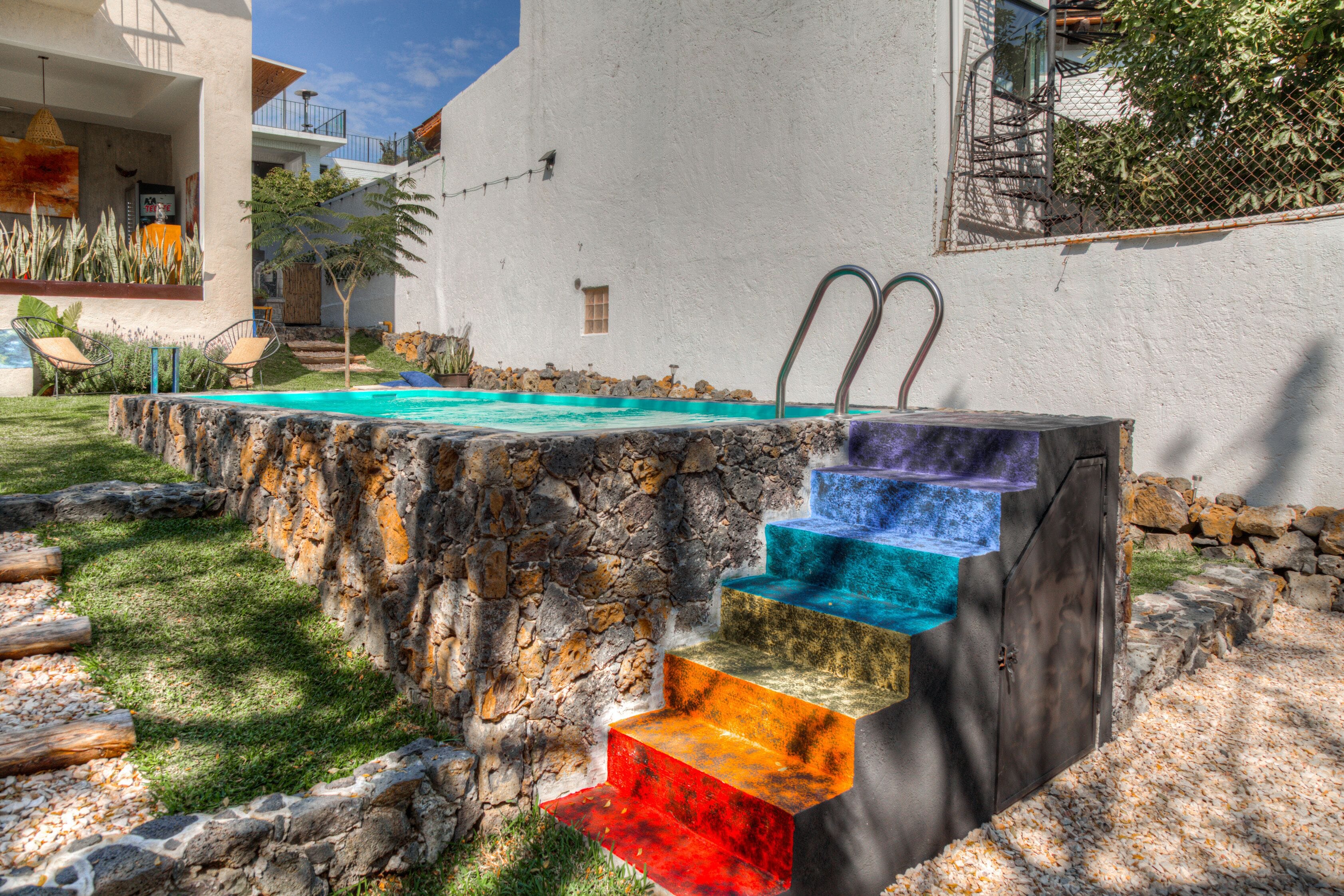 Una piscina con una serie de escalones coloridos y originales que conducen al agua, rodeada de un muro de piedra y un césped.
