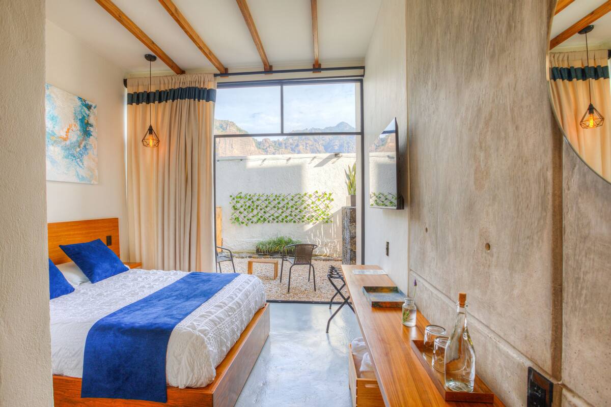 Ein Schlafzimmer mit einem großen Bett, einem Holztisch, einem Fernseher und Blick auf einen Garten durch ein Fenster.