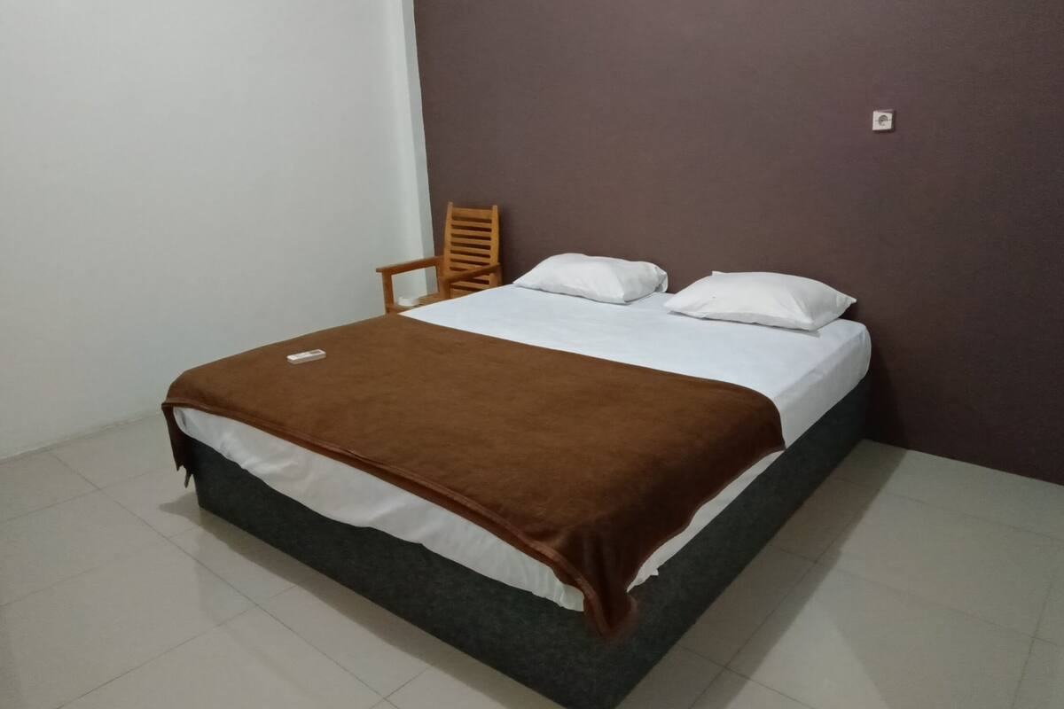 Un lit simple avec une couverture marron, des oreillers blancs et une chaise en bois, dans une pièce aux murs blancs et foncés.