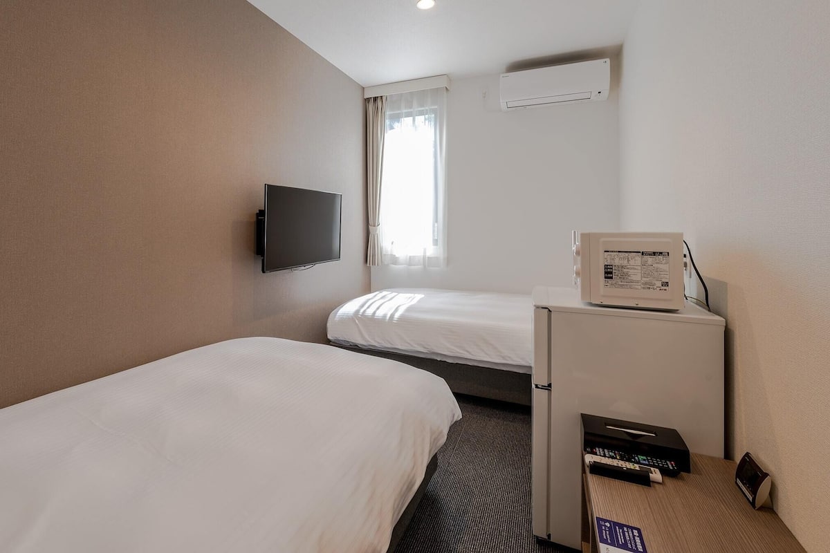 A hotel room with two single beds, a flat-screen TV mounted on the wall, a small refrigerator, and a microwave on a counter.