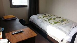 Una habitación pequeña con cama individual, escritorio, silla, cama con colcha de diseño floral y ventana con cortinas. También se observa un teléfono instalado en la pared.