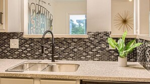 A kitchen with a black and white tiled backsplash, a stainless steel sink, and a decorative wall with a 'Greetings From Me' sign.