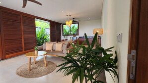 A modern living room with a sofa, a coffee table, and a TV. There are large windows with plants outside. - Sumberkima Resort (Pemuteran)