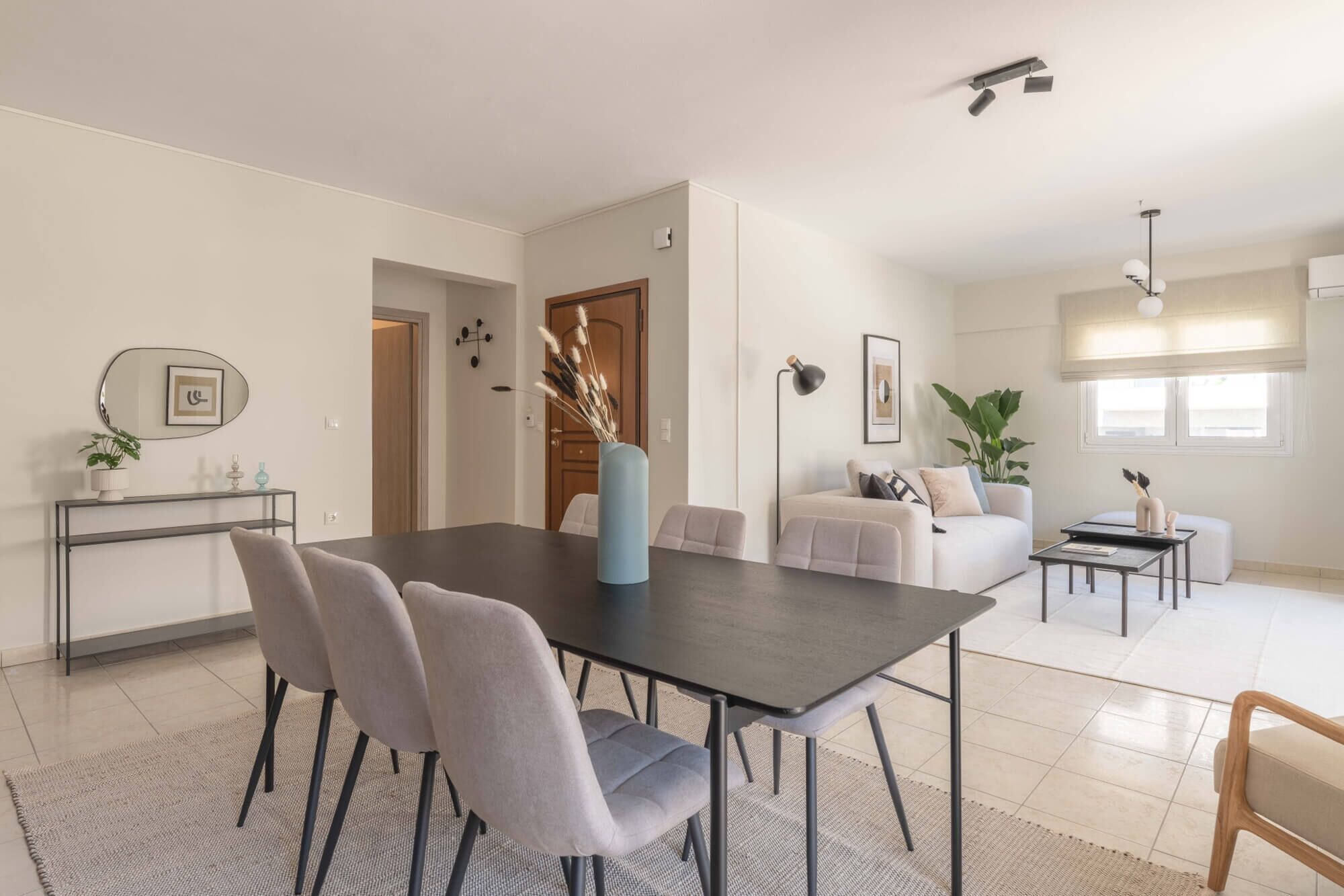 Un salon moderne avec une table à manger en bois foncé, des chaises à revêtement gris, un canapé blanc et une petite table basse. On aperçoit une plante en pot sur une console et une grande fenêtre donnant sur un beau paysage.